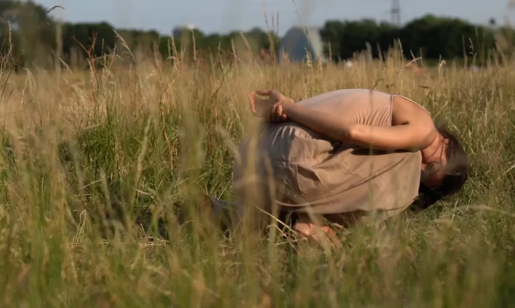 It's summer in Hackney Marshes. The grass is yellow and dry. Claire is curling up onto herself like a shell or a snail, her hands behind her back. She seems deep relaxed and embedded in the place. She has become fully part of the landscape.