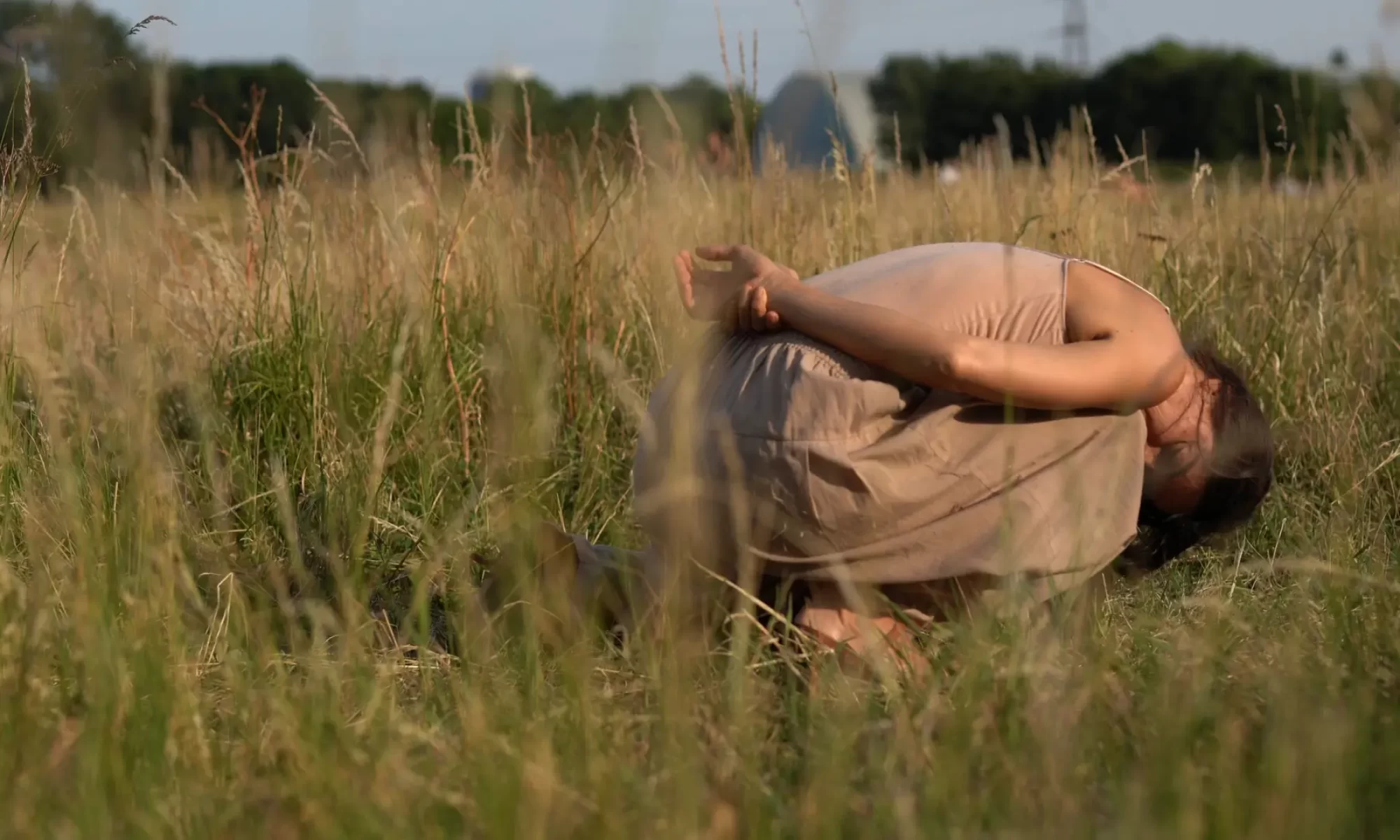 It's summer in Hackney Marshes. The grass is yellow and dry. Claire is curling up onto herself like a shell or a snail, her hands behind her back. She seems deep relaxed and embedded in the place. She has become fully part of the landscape.