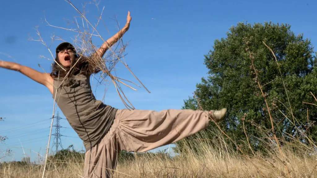 Claire is making a star shape with her right leg standing on the ground and the left at a perpendicular position off the ground. Her mouth is wide open and one of her harm carries dried cow parsley all other. She's standing in the middle of Hackney Marshes at the end of the summer when the grass has turned yellow and dry.