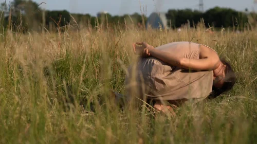 It's summer in Hackney Marshes. The grass is yellow and dry. Claire is curling up onto herself like a shell or a snail, her hands behind her back. She seems deep relaxed and embedded in the place. She has become fully part of the landscape.